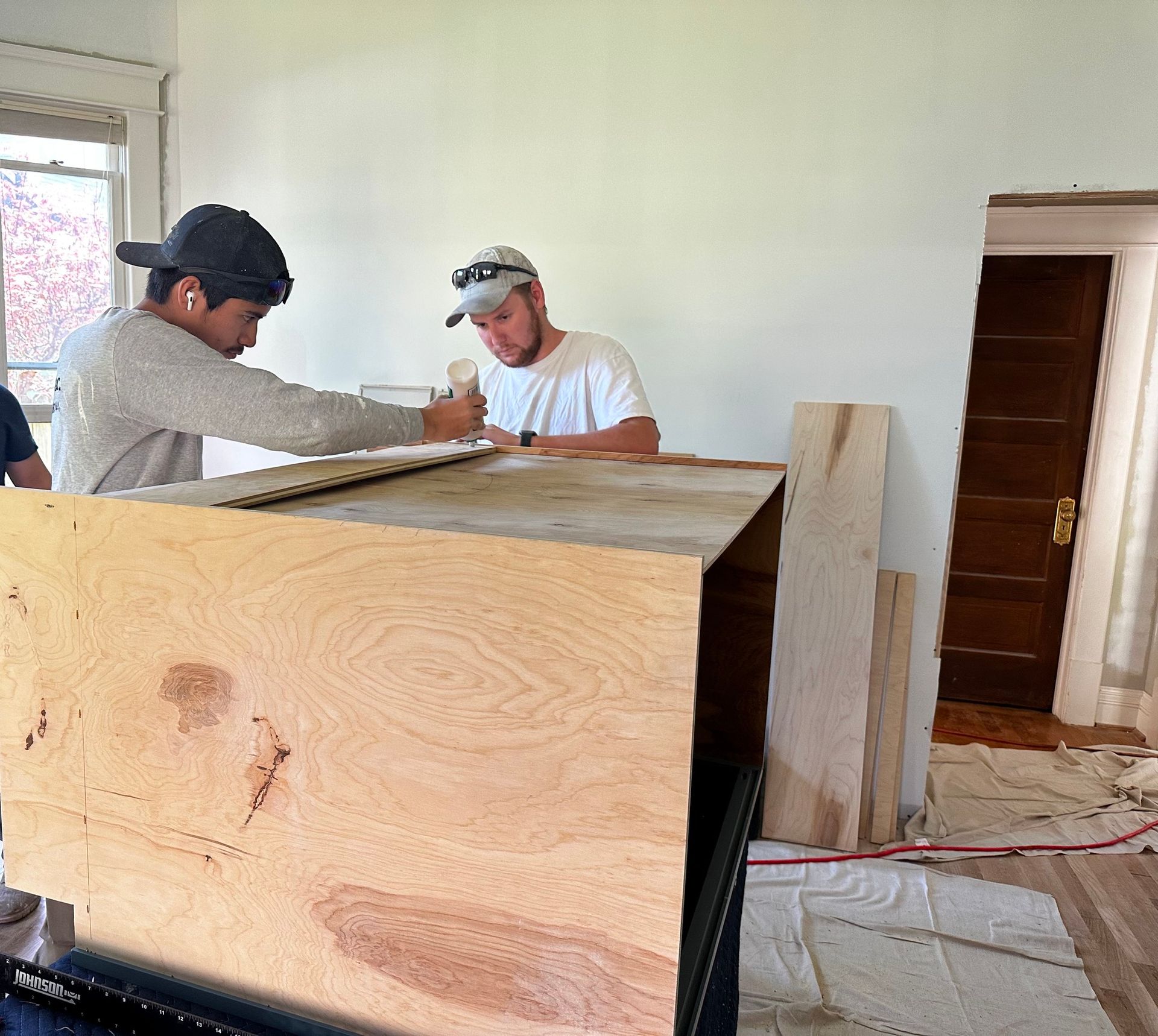 Two men building a wooden cabinet in a room with white walls and a wooden door.