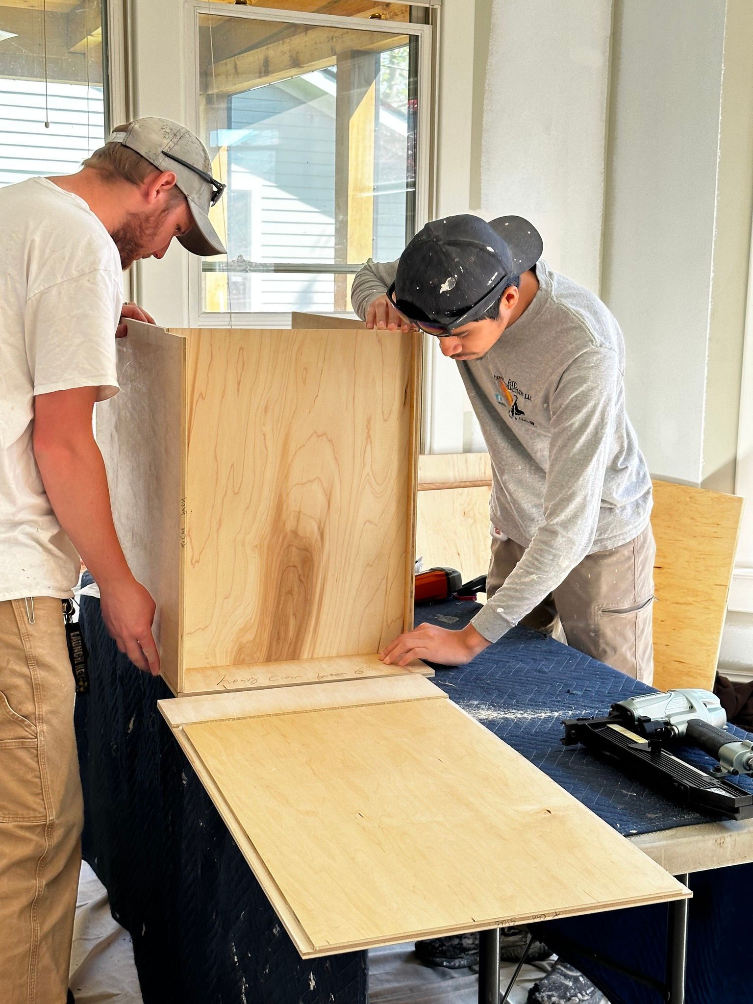 Two people assemble a wooden cabinet indoors, working on a countertop with tools nearby.