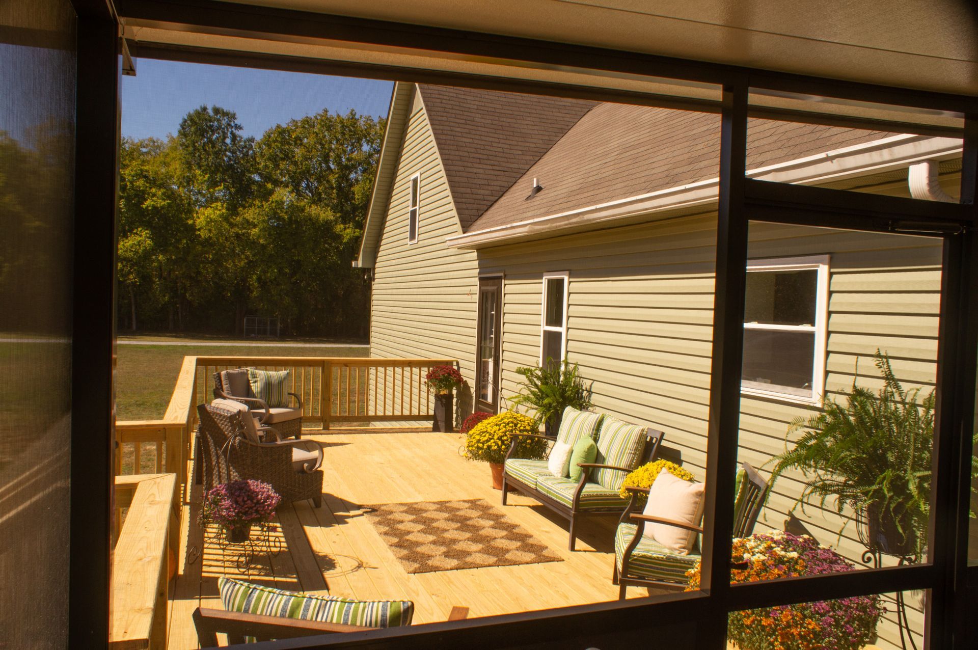 View from a screened-in porch looking out onto a wooden deck with seating, next to a green house.