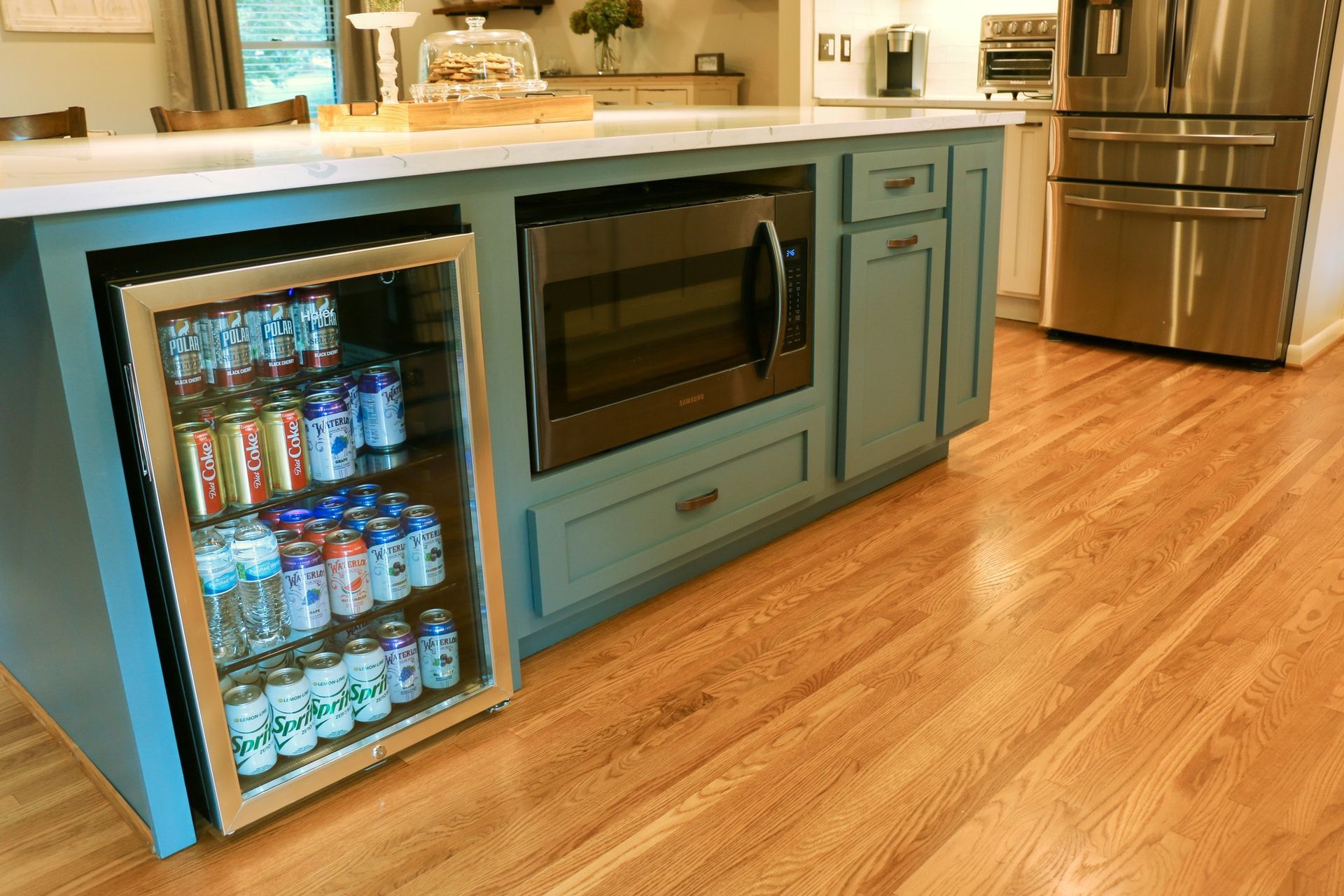 Kitchen island with microwave, beverage cooler filled with cans, blue cabinets, and wood floor.