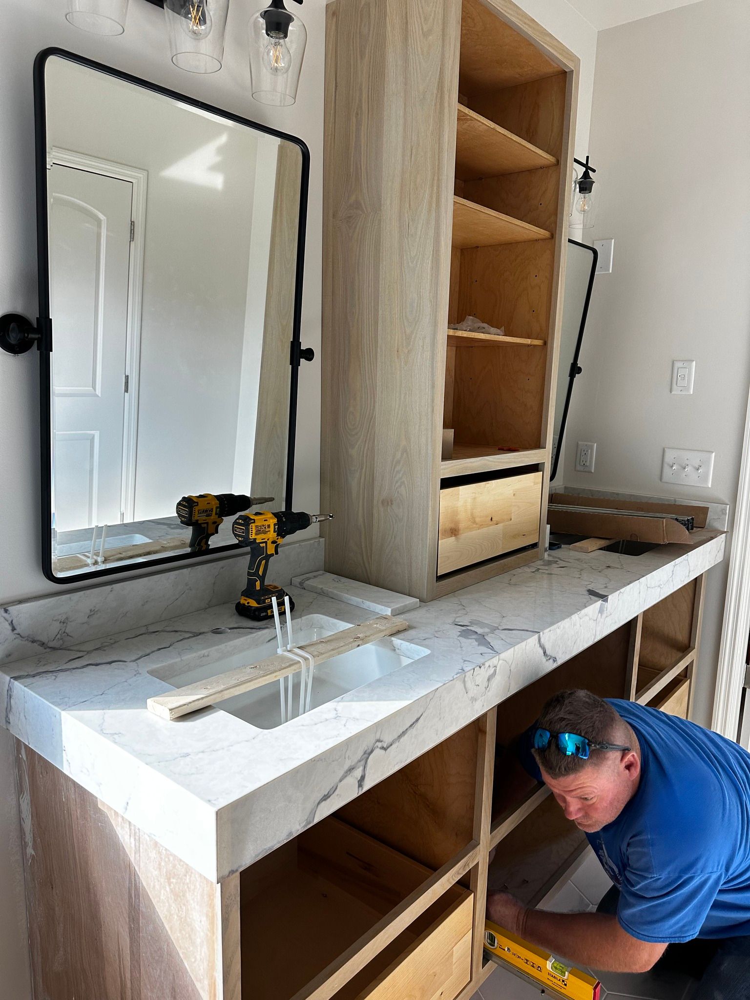 Man installing cabinet drawer in bathroom, light wood cabinets, marble countertop, mirror, and power tools.