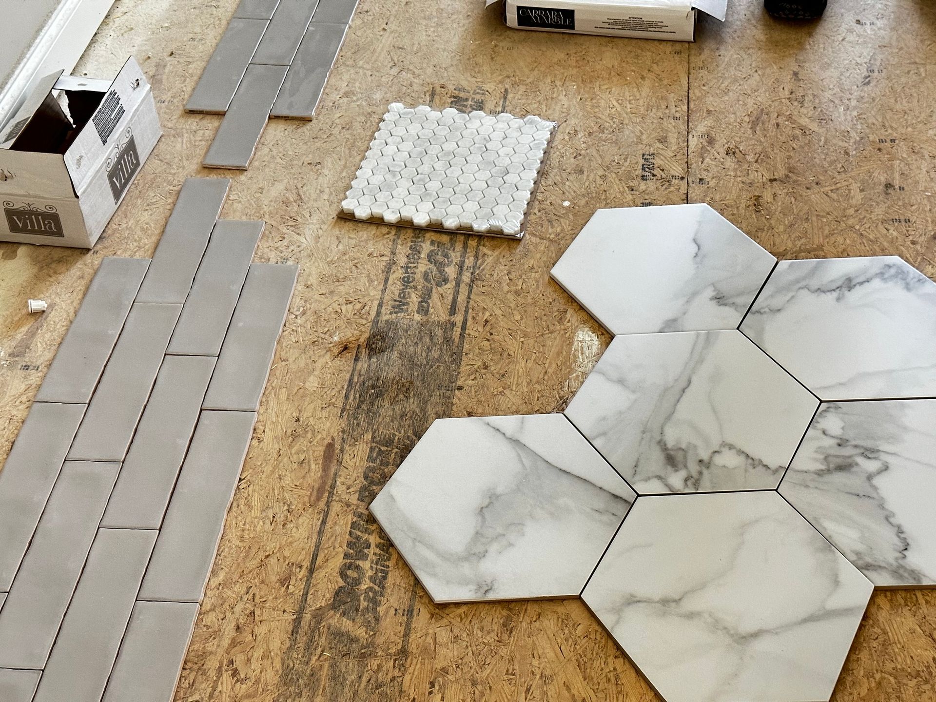 Floor tiling in progress: hexagonal marble-look tiles, gray subway tile, and a small white mosaic set on plywood.