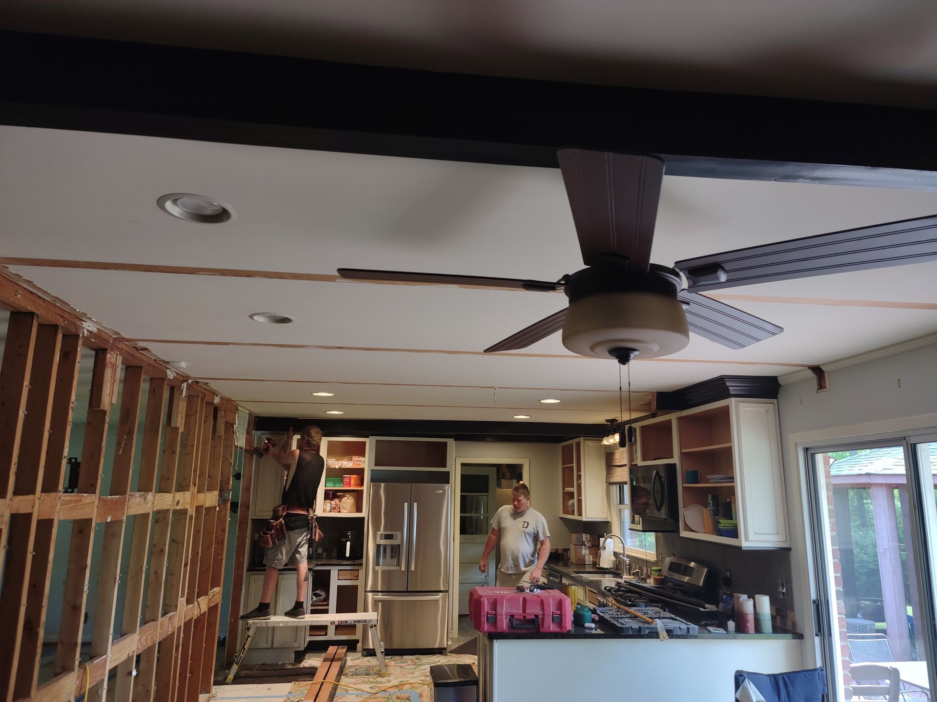 Men renovating a kitchen. One on a ladder, other working by fridge. Exposed wall studs, ceiling fan.