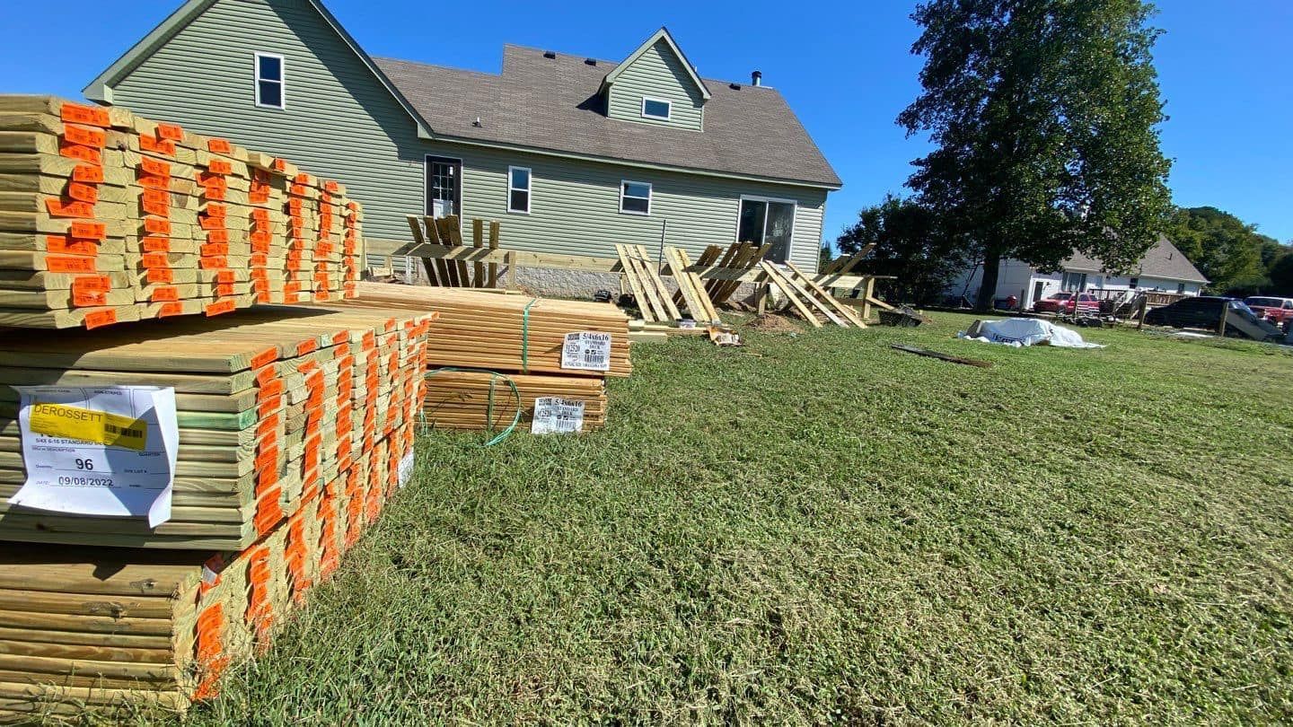 Stacks of lumber sit on a grassy lawn near a green house with a brown roof; construction site.