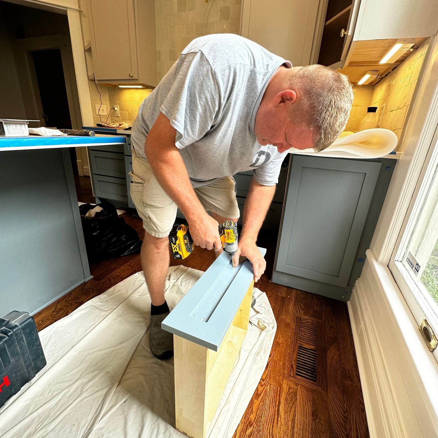 Man drilling cabinet door in kitchen with blue cabinets and a window.