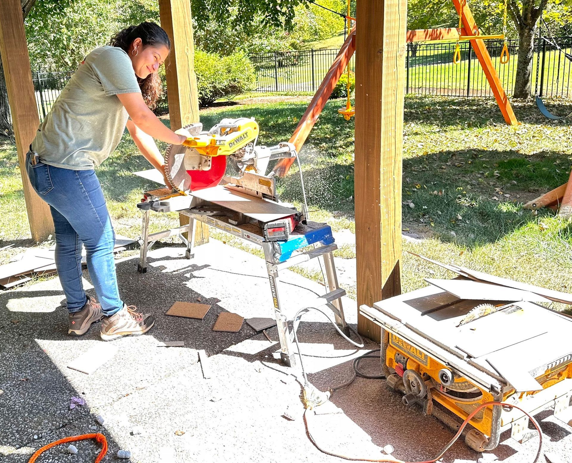 Woman using a miter saw outdoors under a covered structure. She's cutting wood on a saw table.