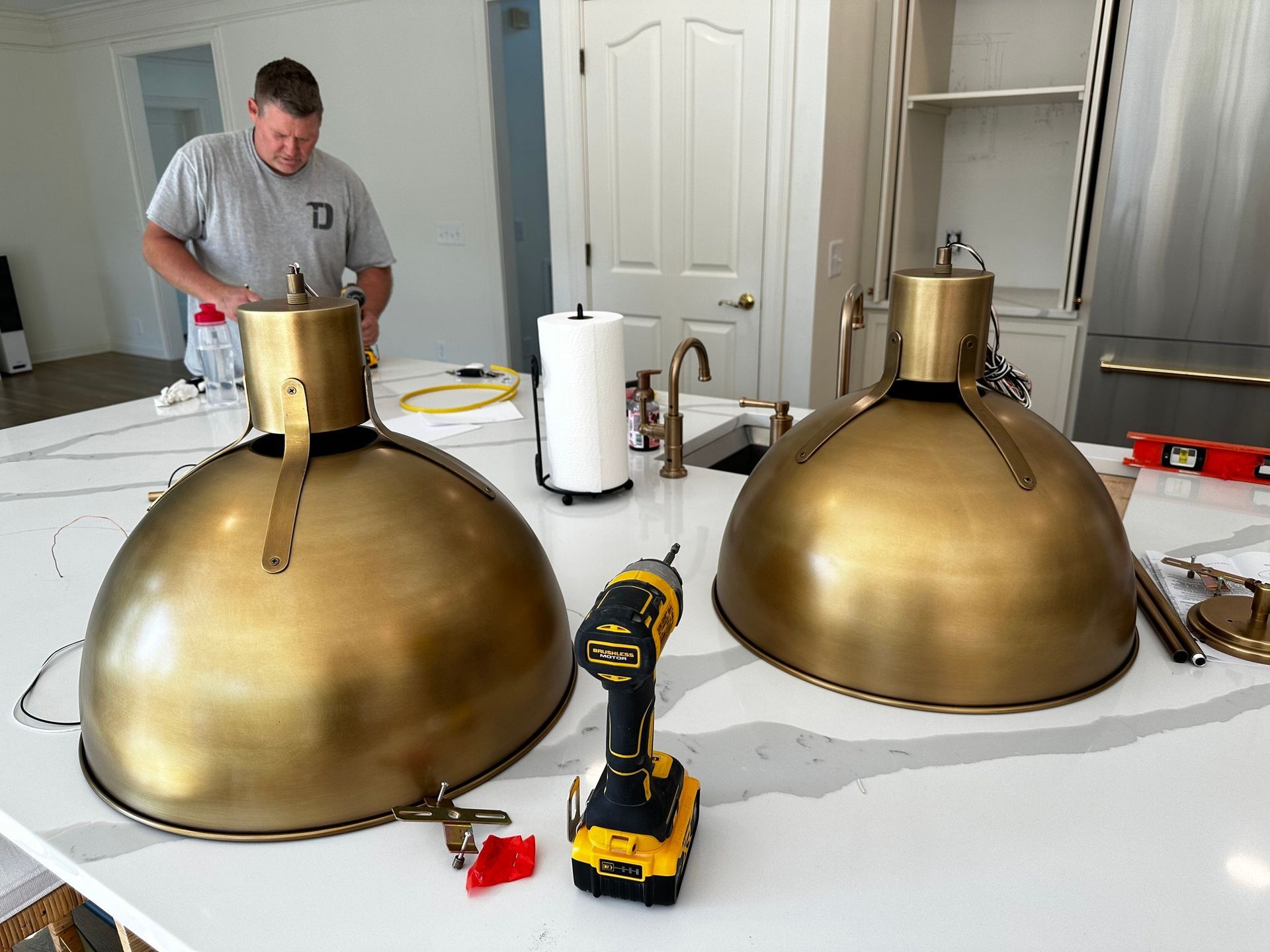 Man installing brass pendant lights over a kitchen countertop. Tools and paper towels are present.