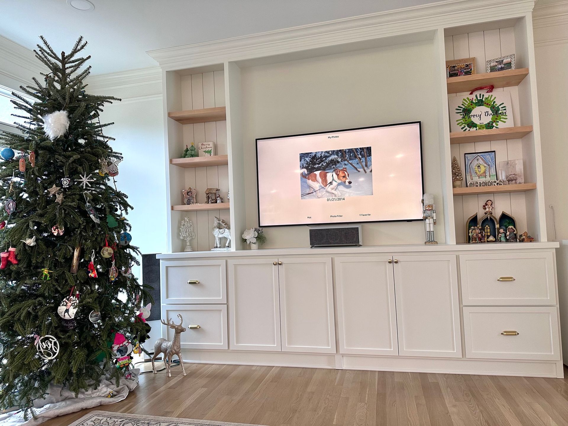A Christmas tree next to a white cabinet with shelves, TV, and decorations in a room with wooden floors.