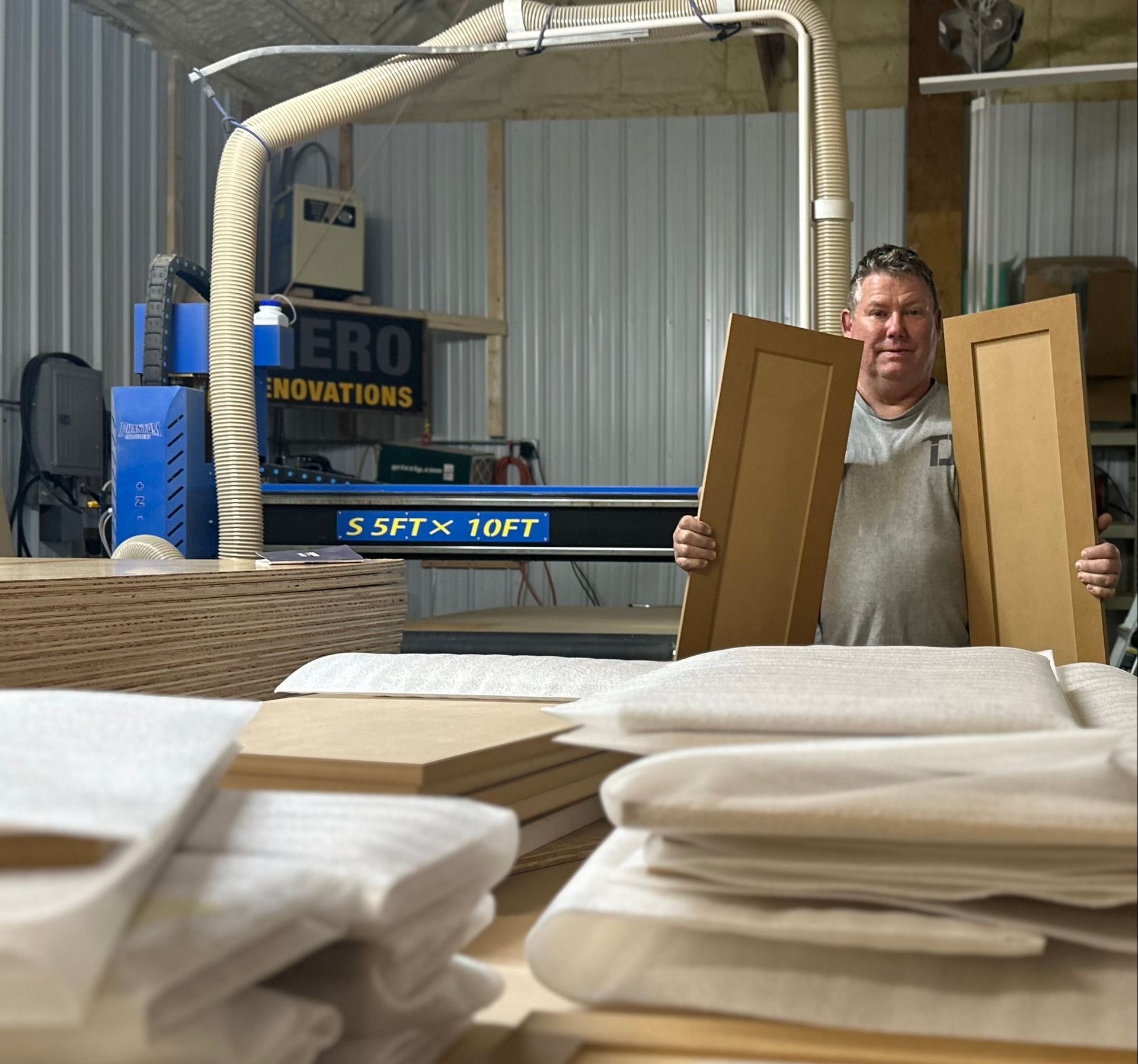 Man holding cabinet doors in workshop with CNC machine and wood.