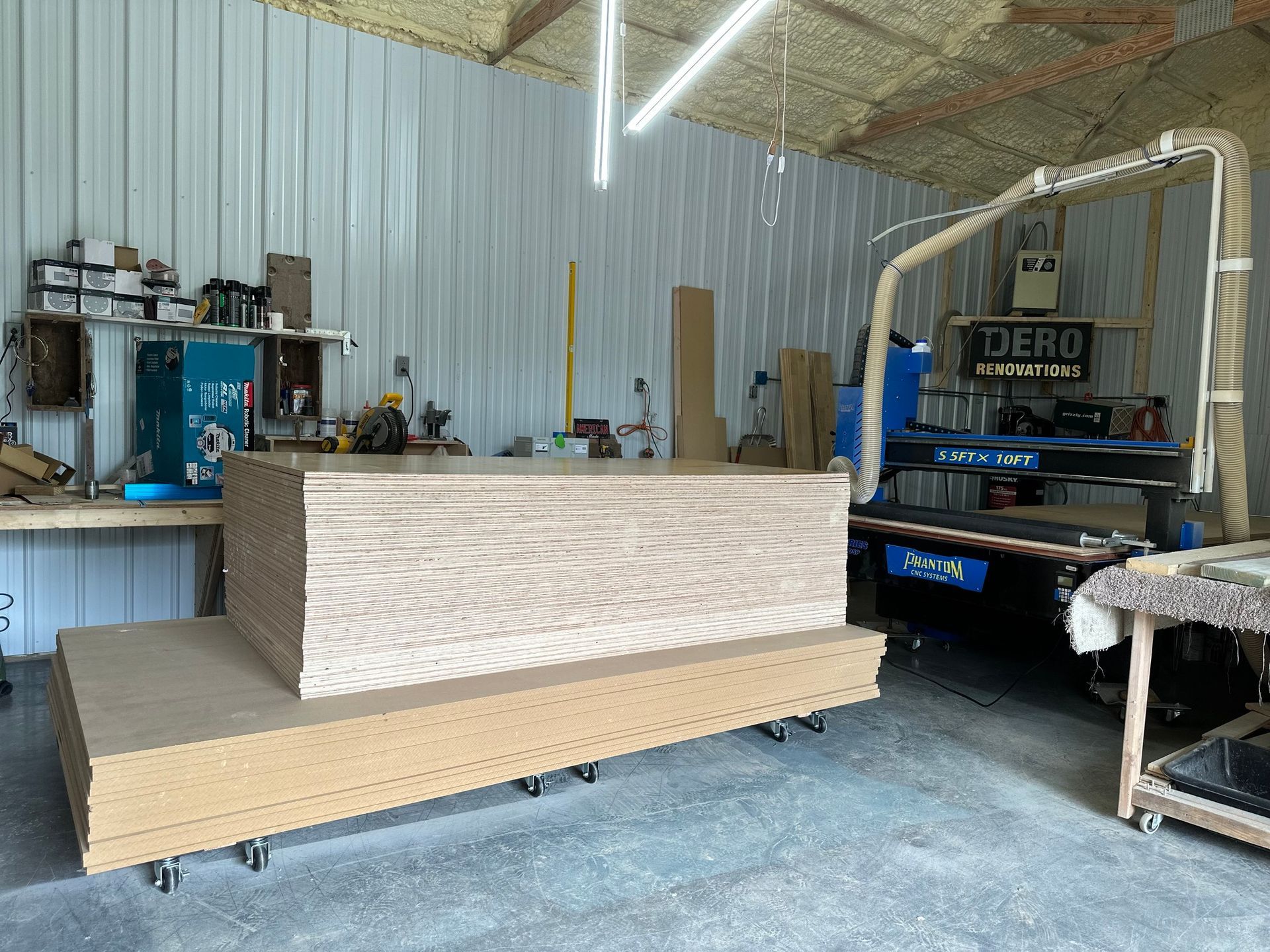 Workshop with stacked wood sheets, CNC machine, tools, and bright lights.