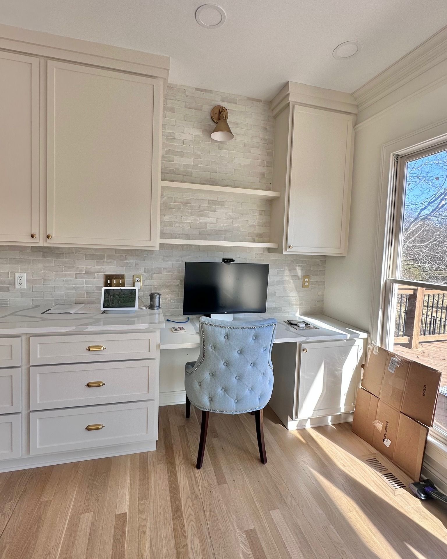 Home office with light blue chair, desk, computer, and white cabinets, with a brick-tiled wall and sunny window.