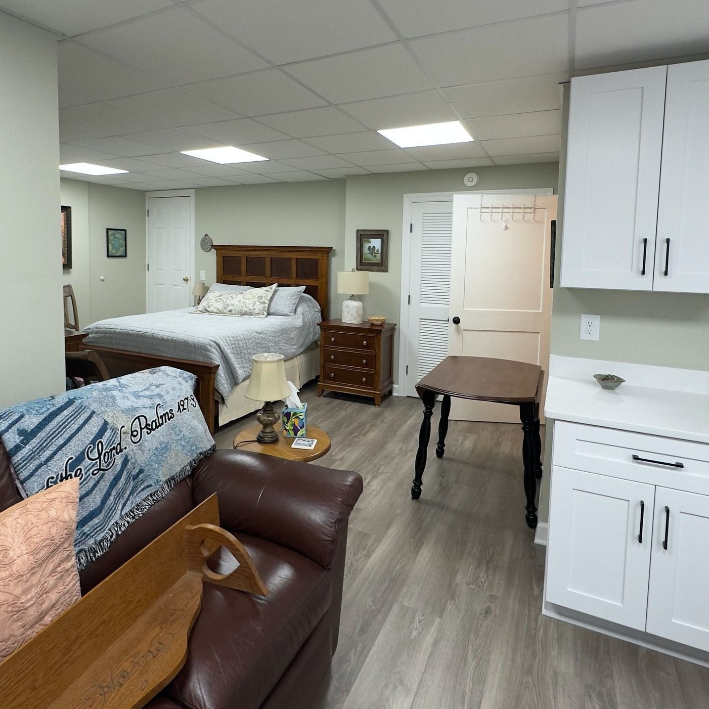 Cozy studio apartment with a bed, couch, kitchen, and dark wood table. Light green walls, white cabinets and doors, and wood floors.