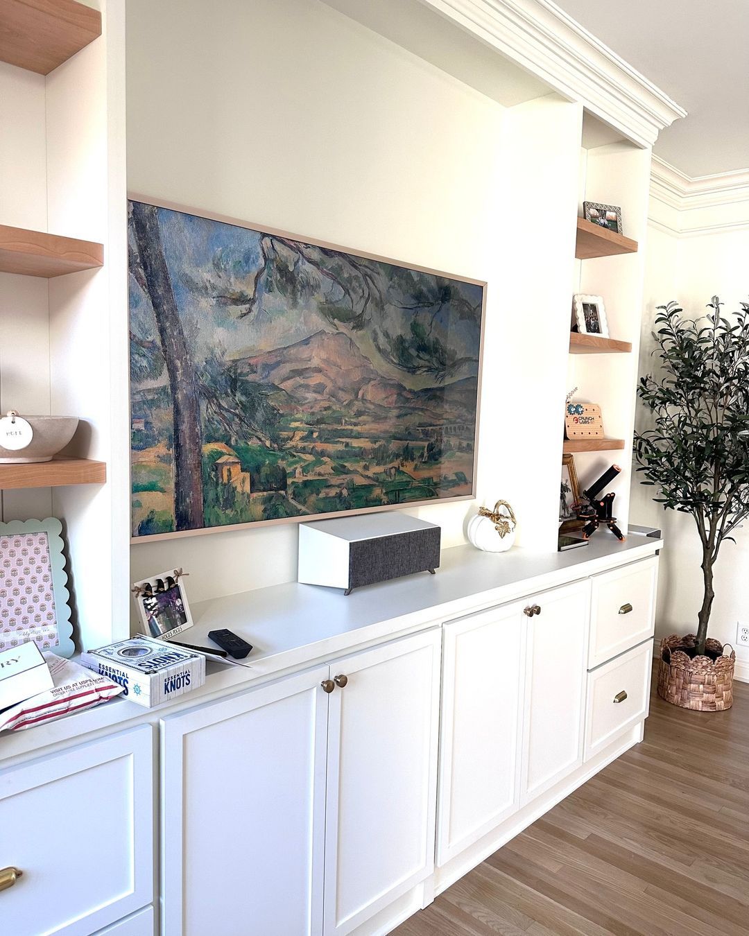 Built-in white cabinets and shelves surround a framed landscape art piece with a soundbar and faux olive tree.