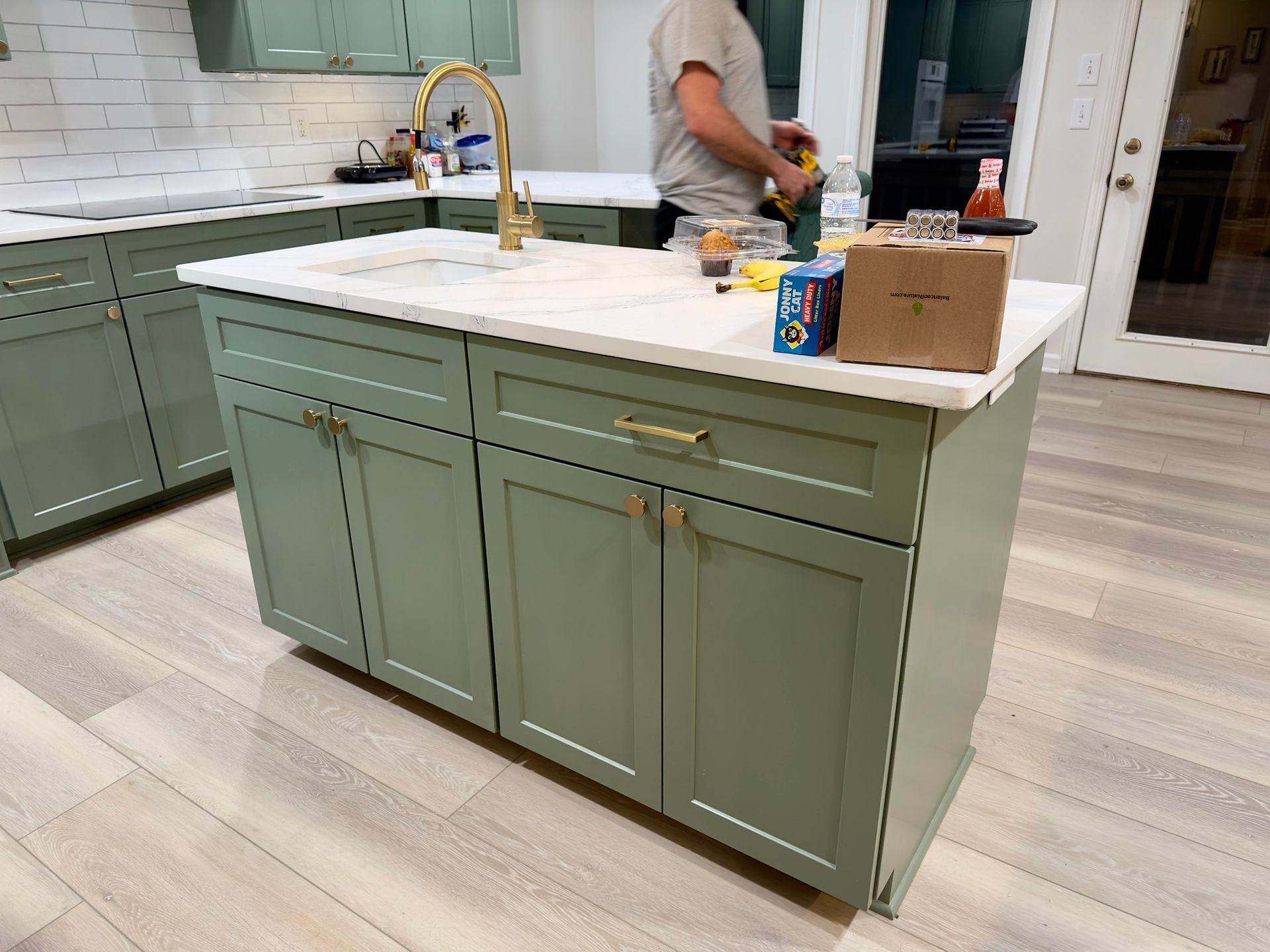 Green kitchen island with white countertop, gold faucet. Person working in the background.