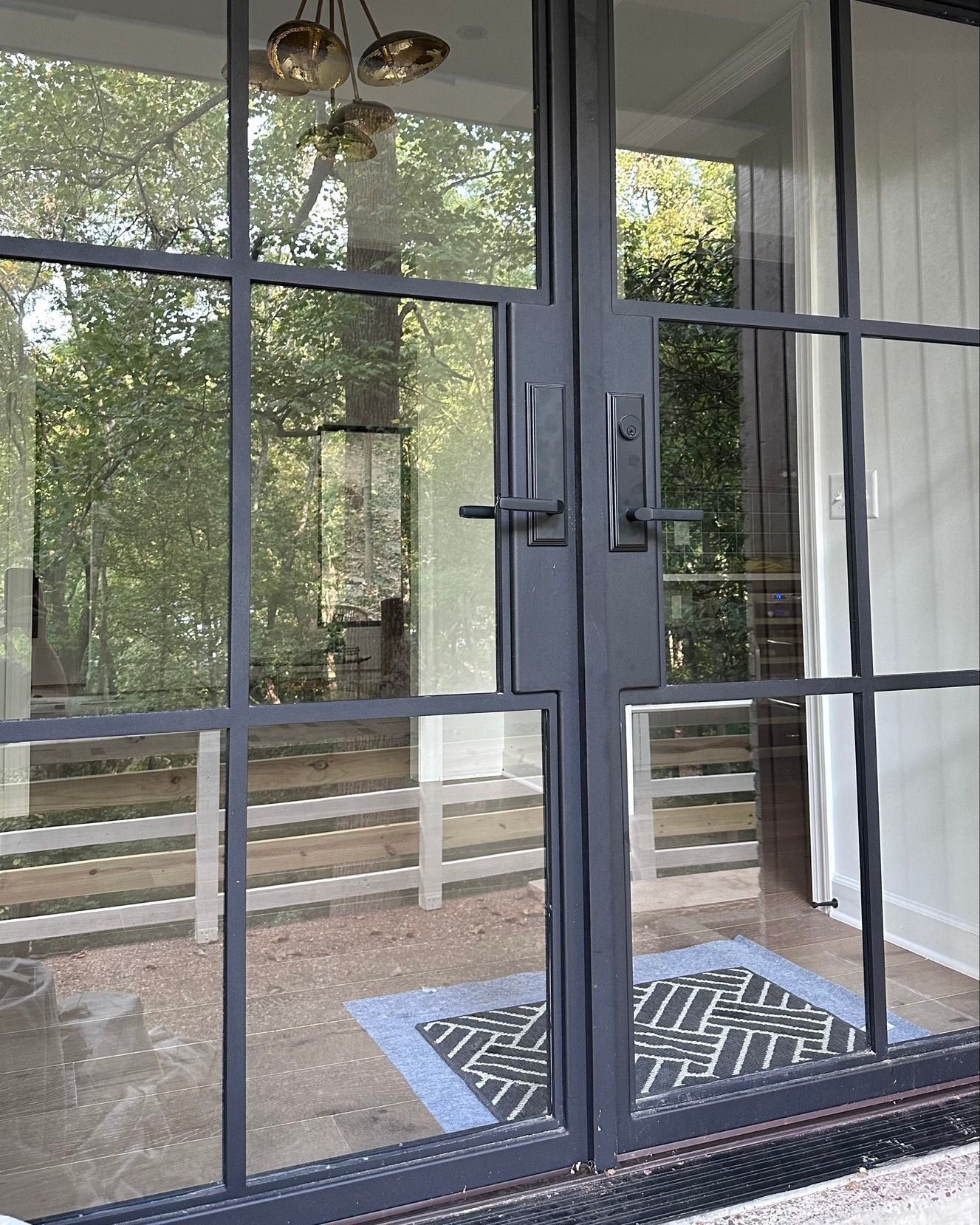 Dark gray steel-framed double doors with handles, looking out onto a patio with a doormat and foliage.