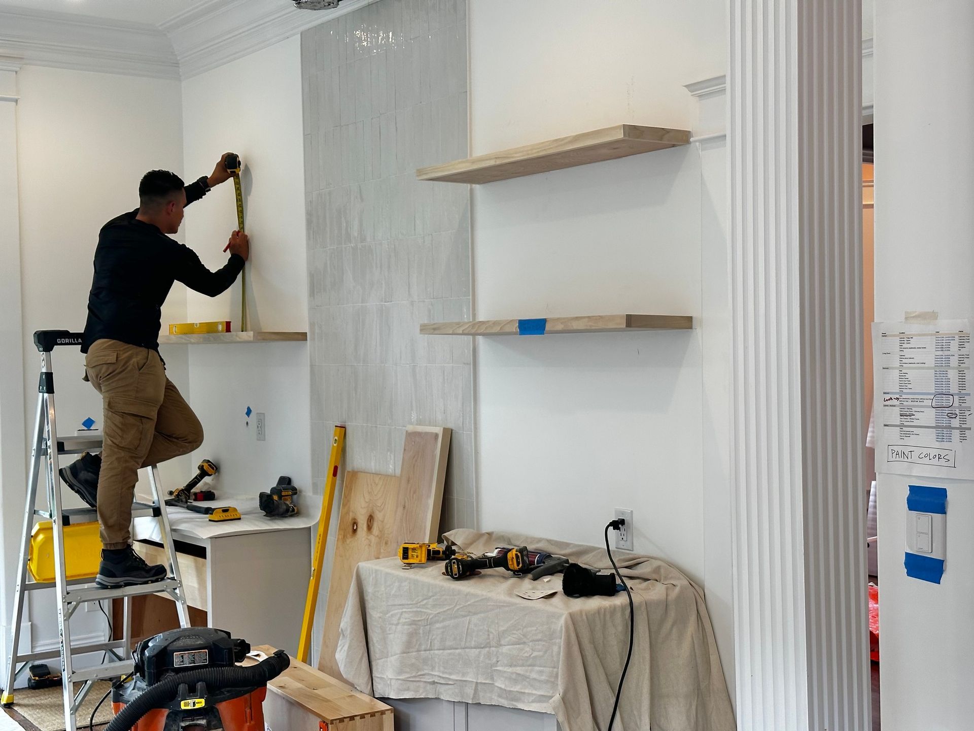 A man on a ladder measures a wall while installing shelves. White walls, tools, and wood are present.