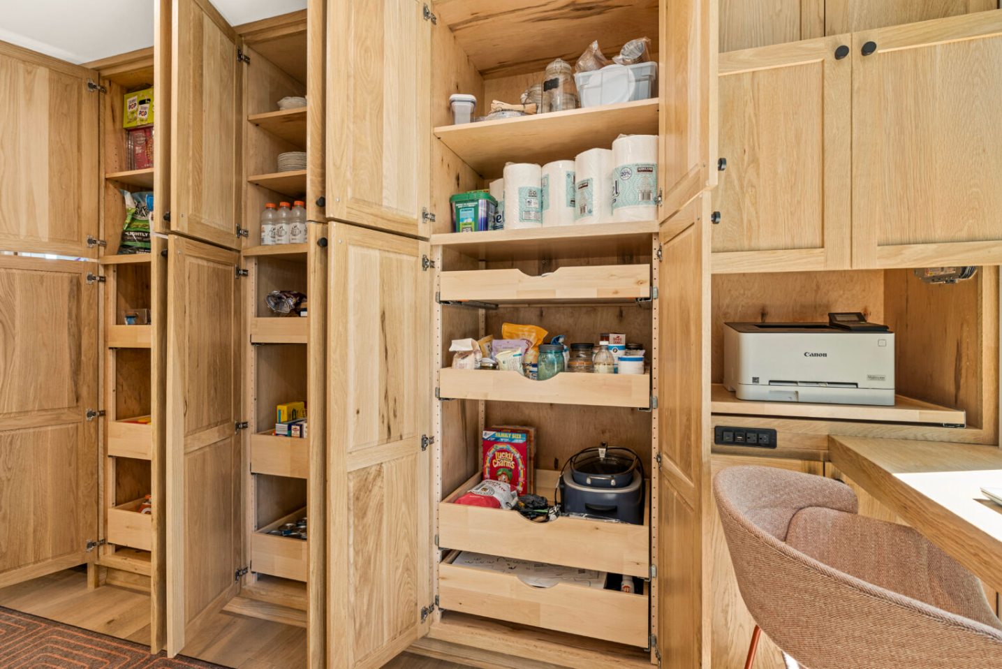 Wooden pantry with pull-out shelves and open doors, next to a built-in countertop with a microwave and cabinets.