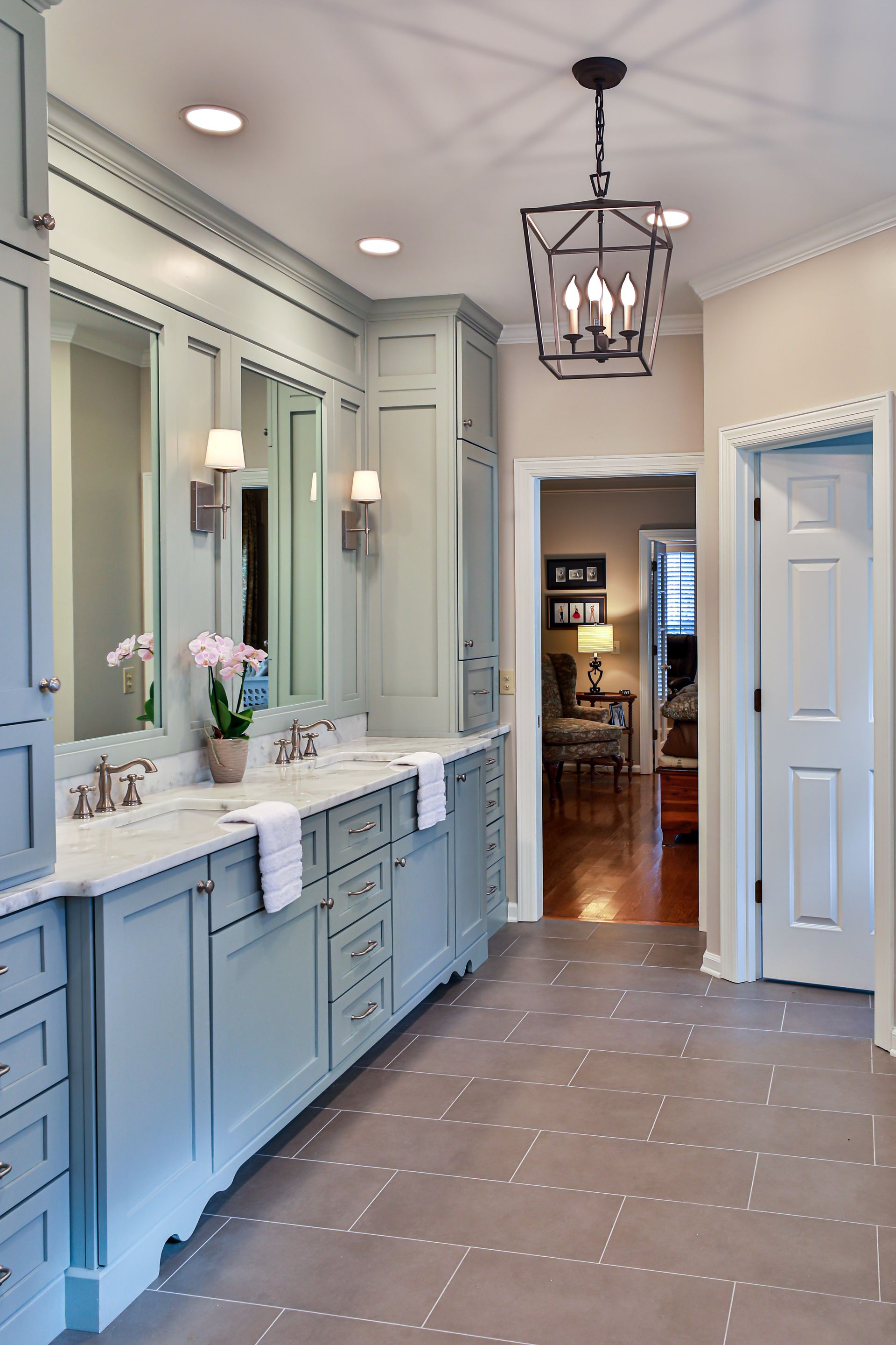 Light blue bathroom with marble countertop, double sinks, mirrors, and chandelier. Doorway leads to another room.