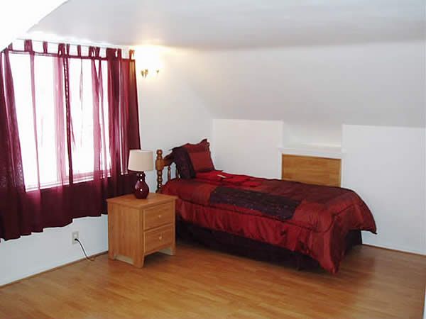Small bedroom with a red bedspread, wooden nightstand, and window with burgundy curtains on a slanted ceiling.