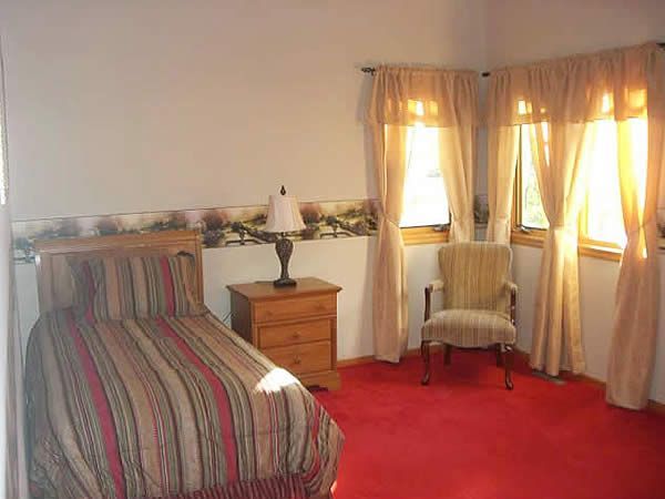 Small bedroom with striped bedspread, red carpet, armchair, dresser, and curtained windows