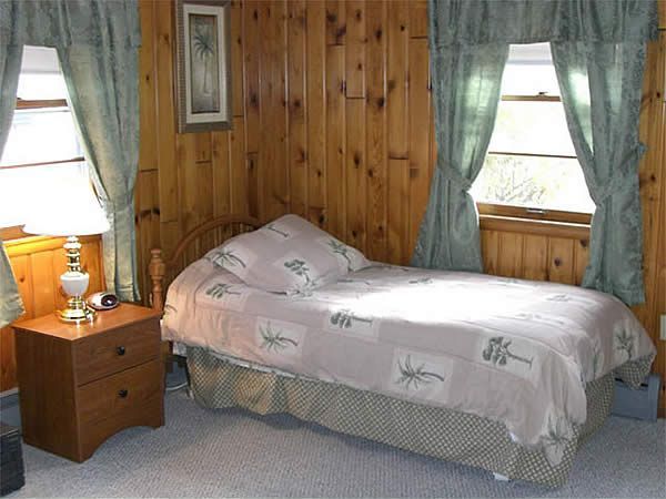 Small bedroom with a single bed, wood-paneled walls, green curtains, and a bedside table with lamp.