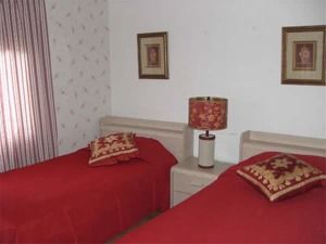 Two red beds in a small bedroom with a white nightstand and floral lamp между them.