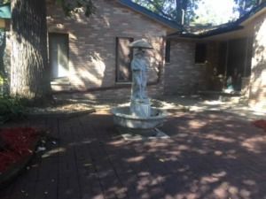 Small stone fountain in a shaded brick courtyard beside a house, with trees and a driveway.