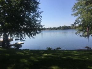 Shaded lakeside park with calm water, trees framing the shore, and a bench near the left bank.