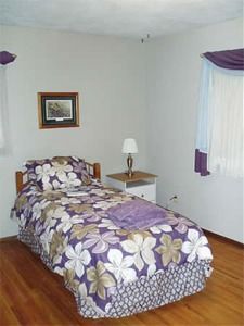 Bedroom with twin bed, purple floral bedding, wooden floor, nightstand, lamp, and framed wall art.