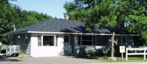 Small white house with a dark roof, front porch, trees, and a sign by the driveway