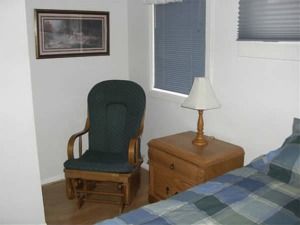 Bedroom with green chair, wooden nightstand, lamp, framed art, and a bed with blue plaid bedding.