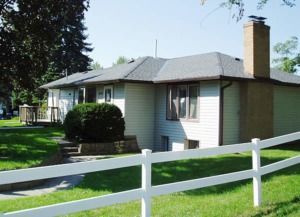 Single-story house with gray roof, white fence, and green lawn in a suburban yard.