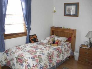 Small bedroom with a floral bedspread, blue curtains, wooden furniture, and a bedside lamp.