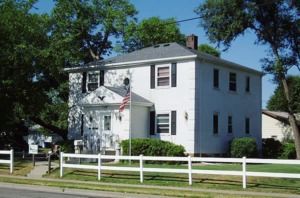 White two-story house with a front porch, American flag, and white fence on a sunny street