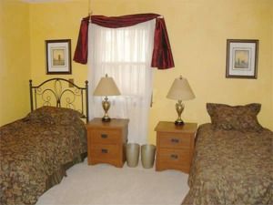 Two twin beds in a small bedroom with a window, nightstands, lamps, and framed pictures on yellow walls