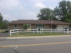 Single-story beige building with a brown roof behind a white fence along a roadside.