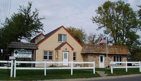Small tan building with red trim and a white fence, with an American flag and trees outside.