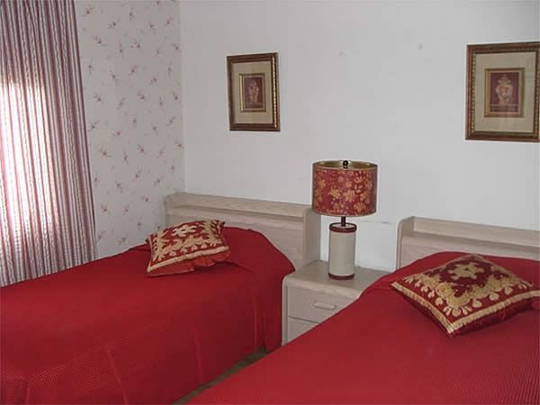 Two red beds with patterned pillows in a small bedroom, separated by a nightstand with a lamp