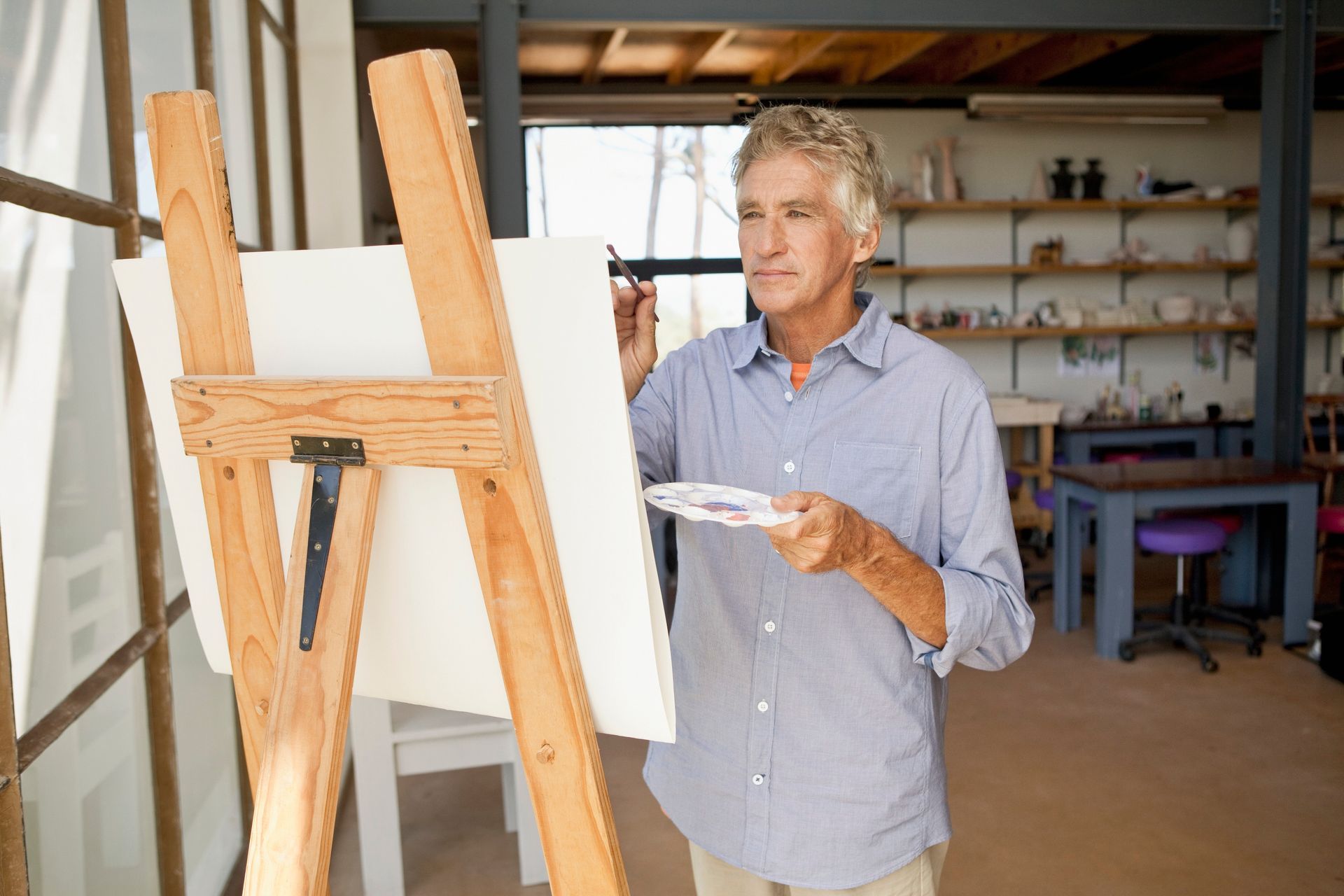 Artist painting at an easel in a workshop, holding a palette knife and facing a blank canvas.