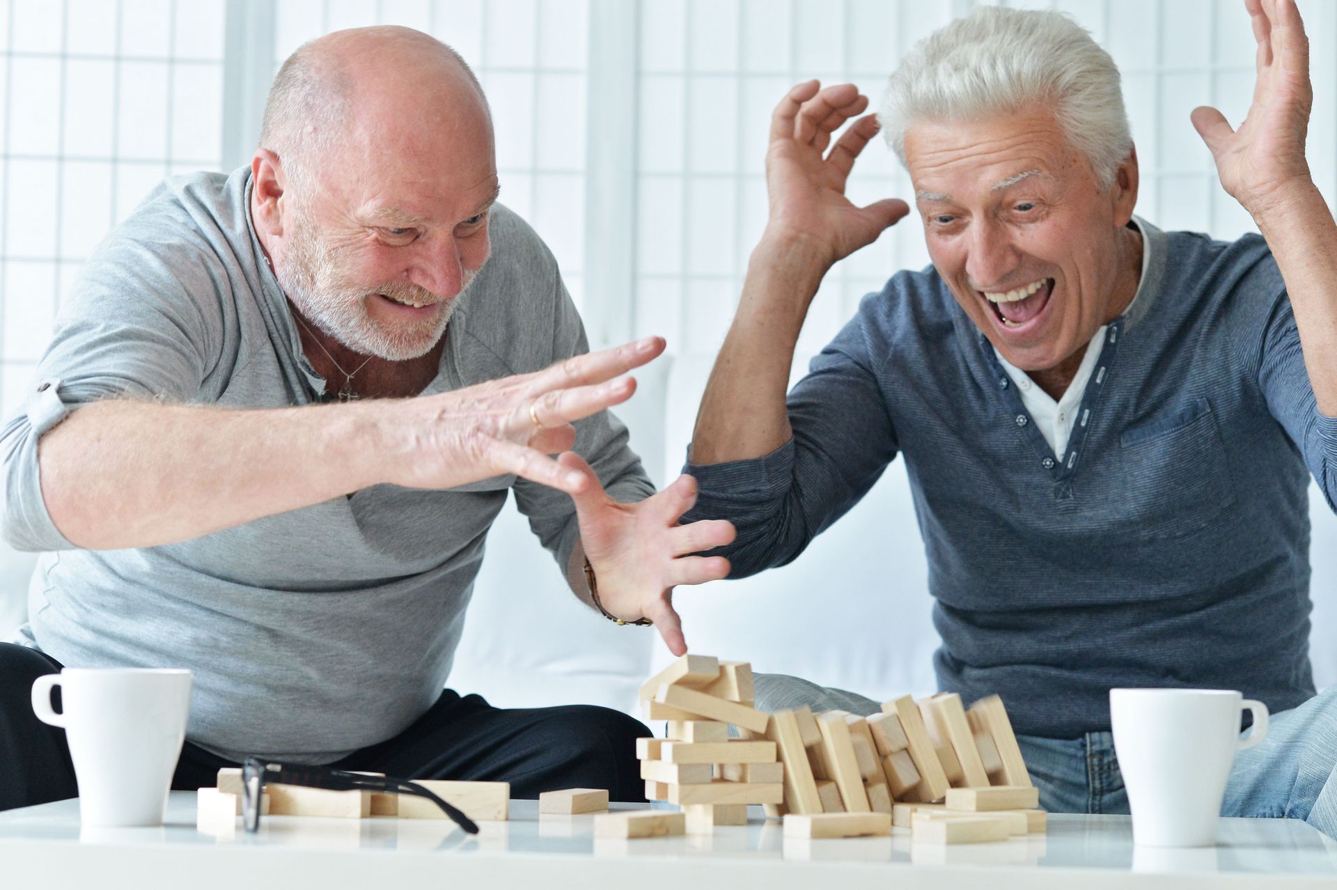 Two smiling people reaching toward a tumbling wooden block tower on a table