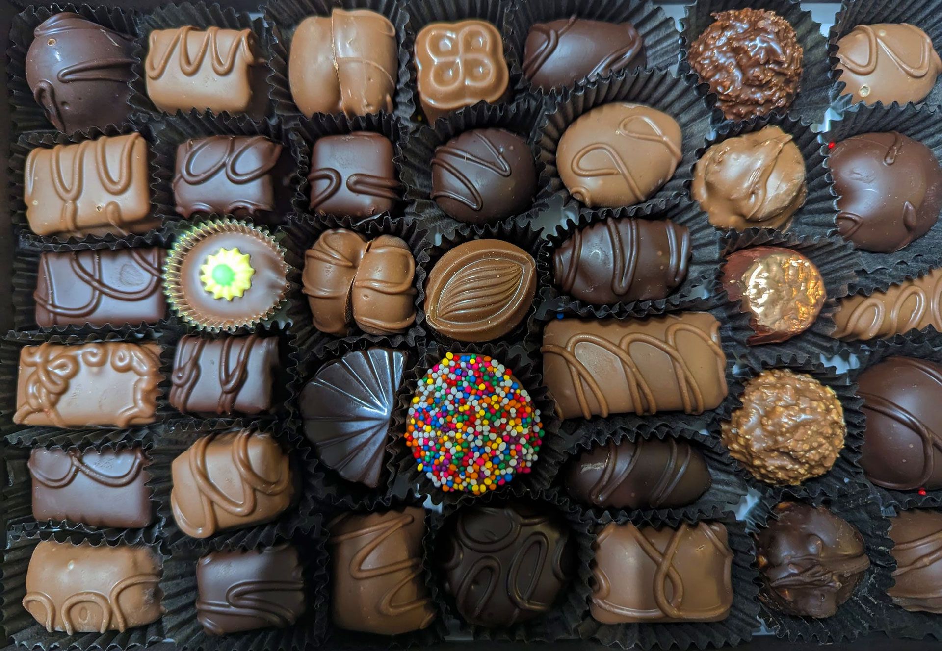 Box of various chocolate candies in black paper cups.