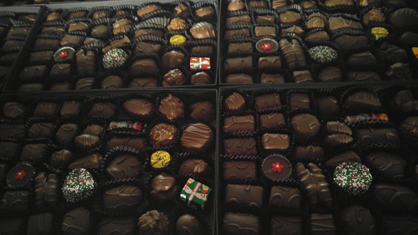 Box of various chocolates, dark brown and golden hues, arranged in rows. Some have toppings, decorative candies.