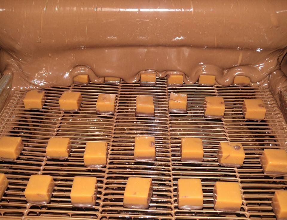 Caramel cubes on a conveyor belt being coated in flowing chocolate.