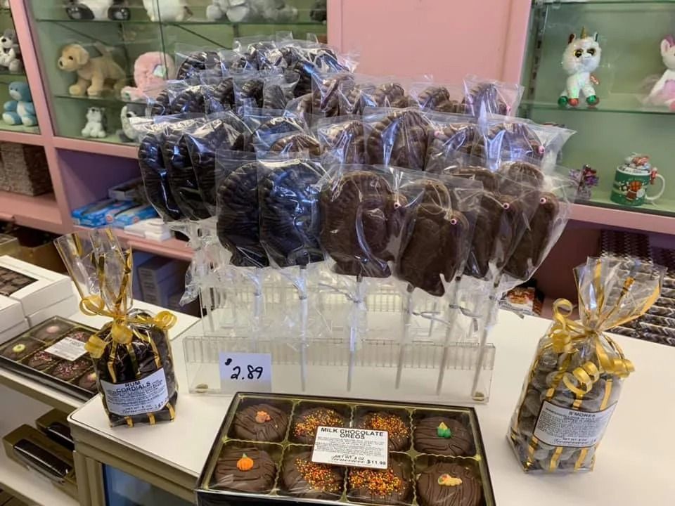 Chocolate treats: lollipops, boxed chocolates, and pretzel bag, displayed in a store with shelves of toys in the background.