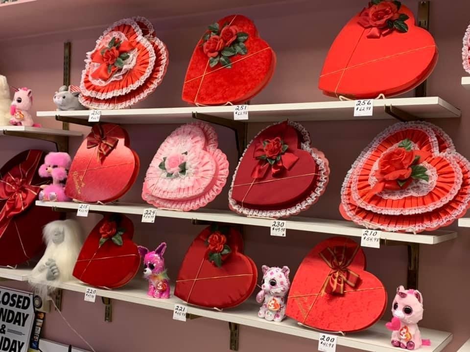 Shelves display heart-shaped boxes of varying shades of red, adorned with bows and flowers, alongside stuffed animals.