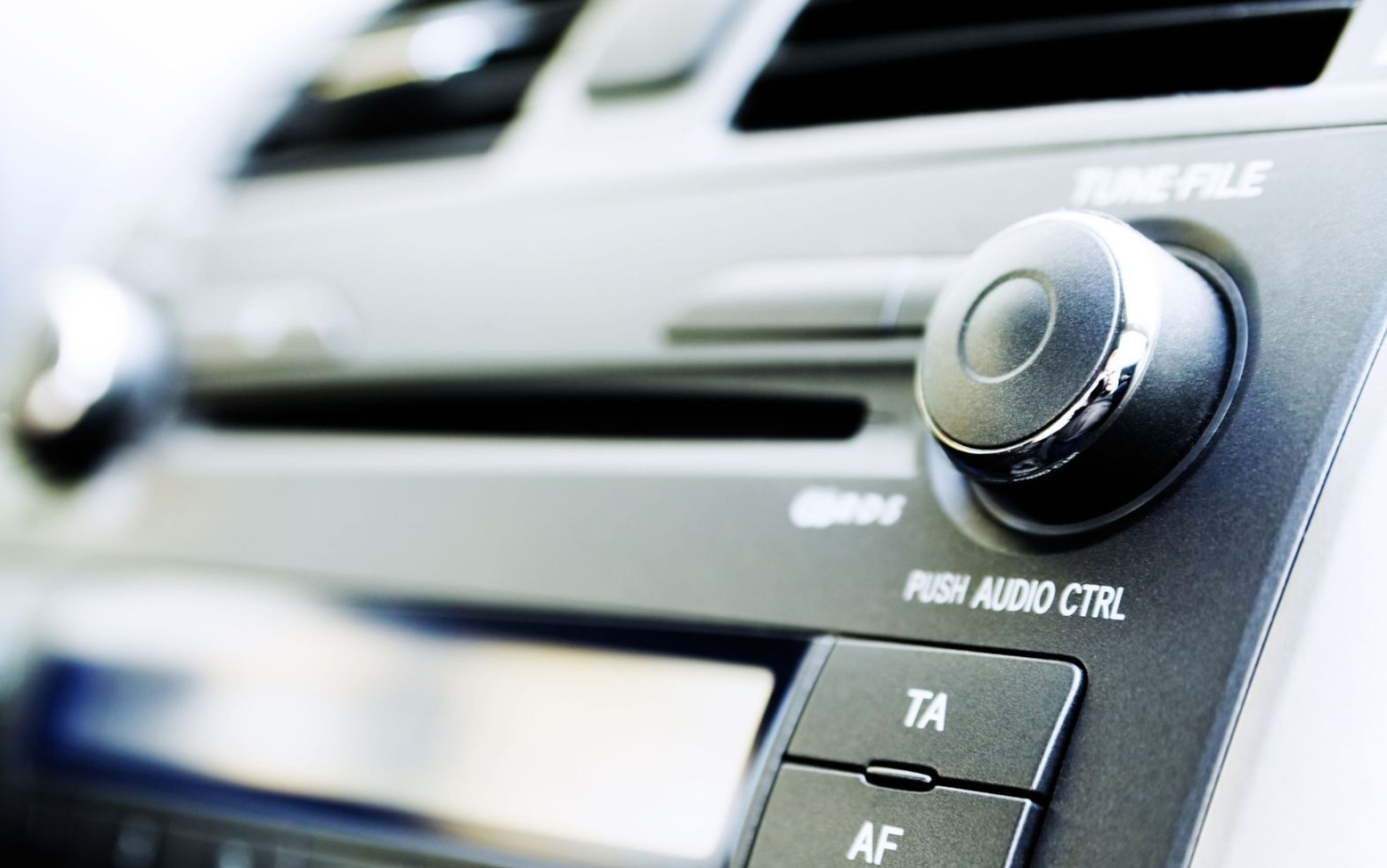 Close-up of a car's dashboard with a radio dial, buttons, and air vents. Gray and black tones.