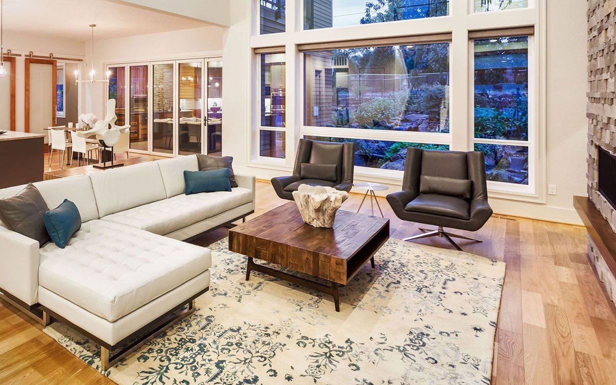 Bright living room with white sectional sofa, dark chairs, large windows, and a wooden coffee table over a patterned rug