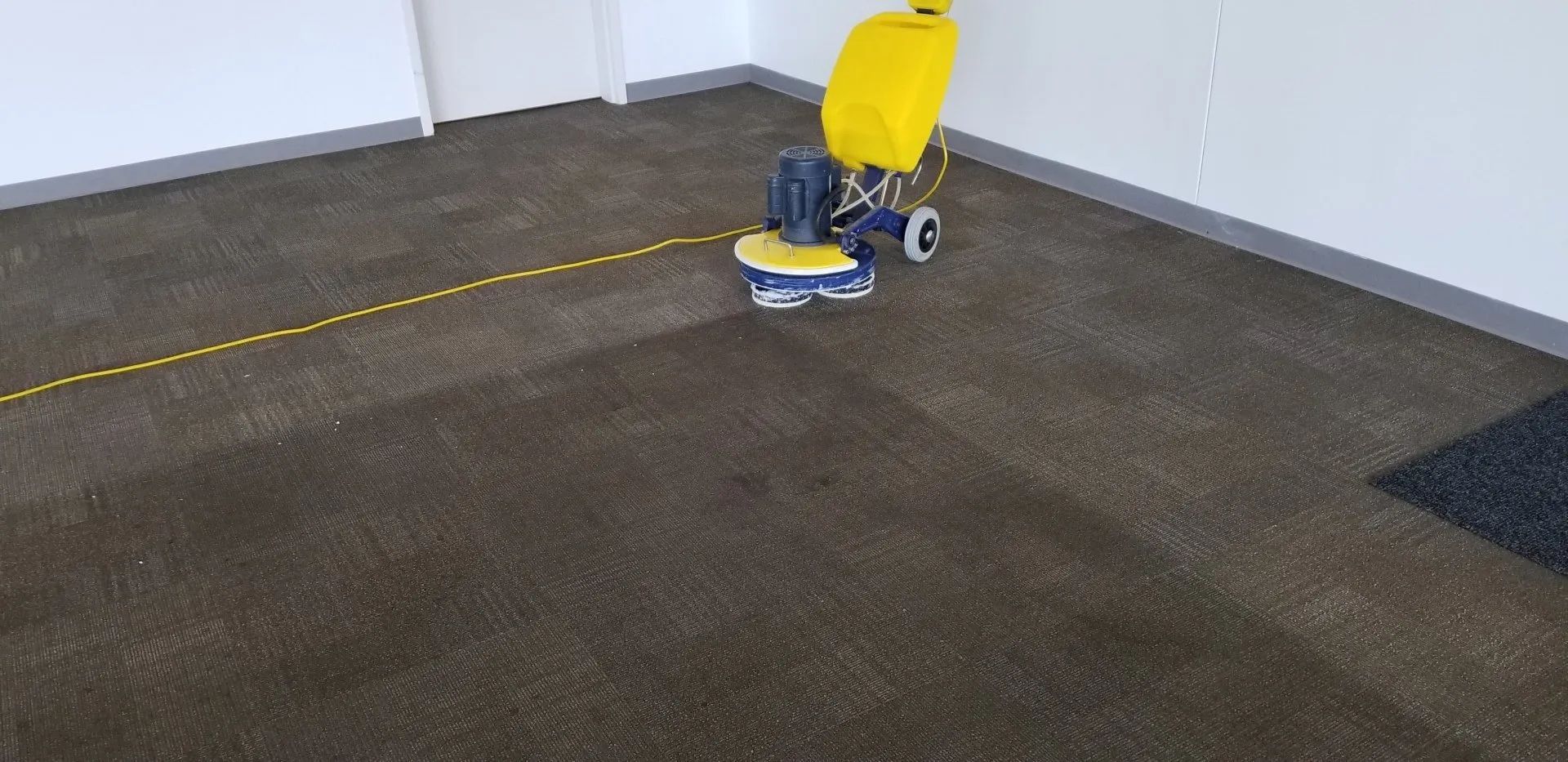 Yellow floor buffer on a dark carpet in an empty room with white walls.