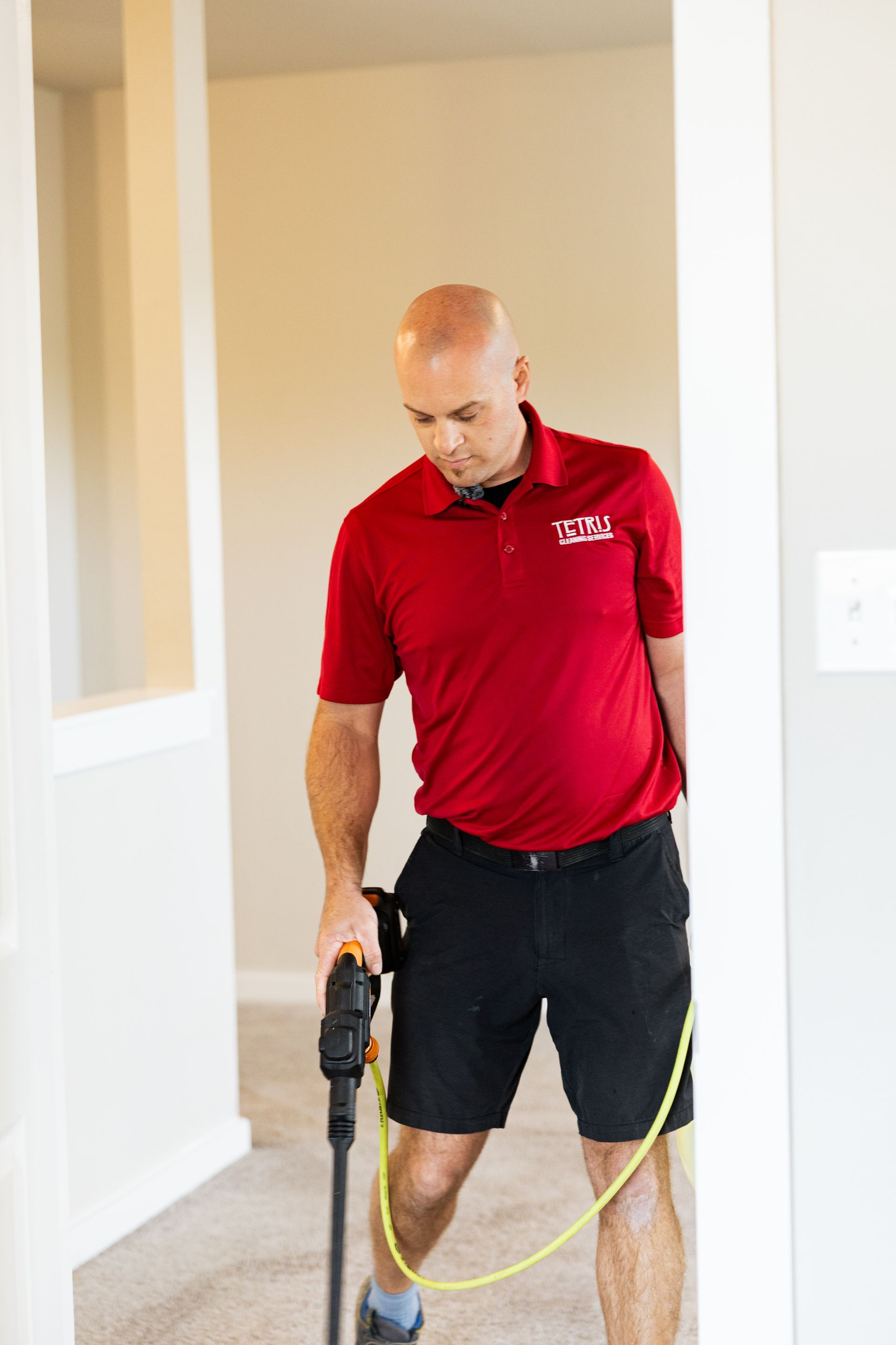 Man in red polo and black shorts holding a power drill, standing indoors by a wall with a yellow cord.
