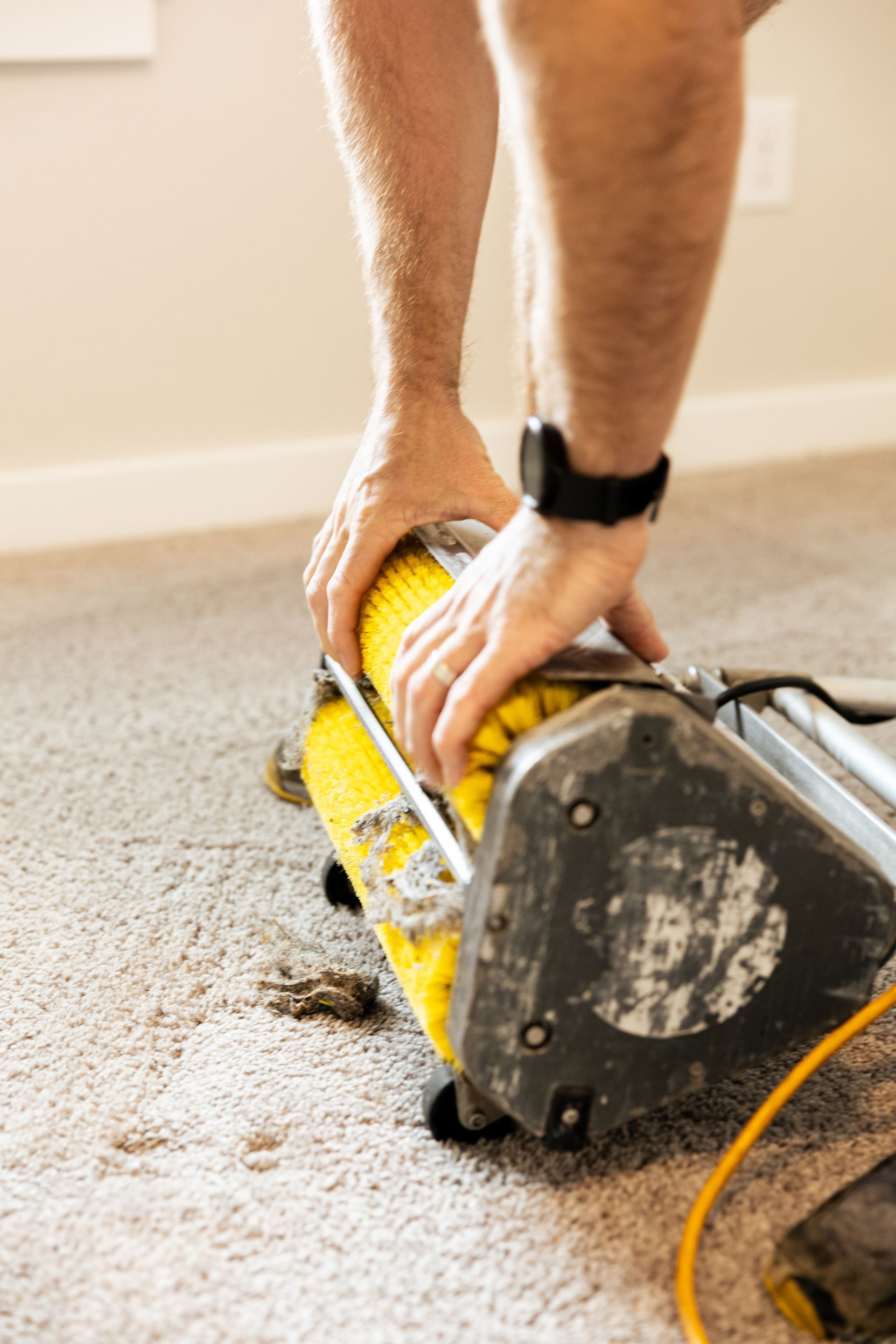 Hands and legs guiding a yellow floor sander on carpeted flooring.